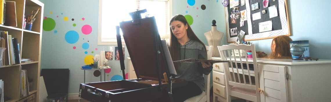 A woman sitting at her easel painting in a colourful room